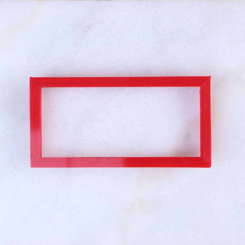 May include: A red plastic frame with a rectangular opening. The frame is on a white surface.