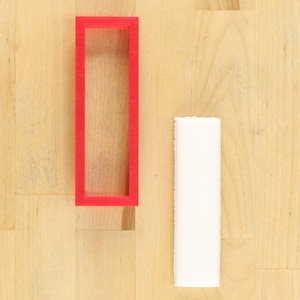 May include: A red plastic frame with a white rectangular object next to it on a wooden surface.