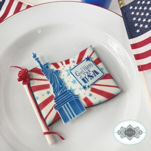 Puede incluir: Una galleta patri&oacute;tica decorada con una Estatua de la Libertad azul y un dise&ntilde;o de bandera con rayas y estrellas rojas y blancas. La galleta tiene el texto "God Bless the USA" y est&aacute; colocada en un plato blanco.