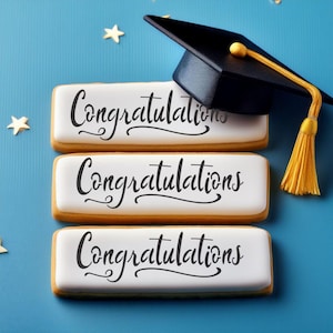 Op de afbeelding: Drie witte rechthoekige koekjes met het woord "Gefeliciteerd" in zwarte cursieve letters op elk koekje. Een zwarte graduatiecap met een gouden kwast ligt op de koekjes.