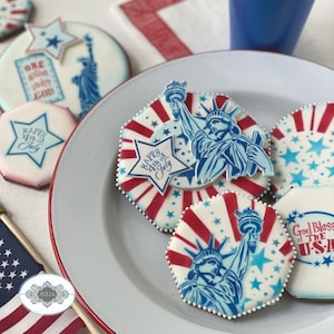Puede incluir: Un plato de galletas decoradas con tem&aacute;tica patri&oacute;tica. Las galletas presentan im&aacute;genes de la Estatua de la Libertad, estrellas y los textos "Happy 4th of July" y "God Bless the USA". Las galletas son blancas con detalles rojos y azules.
