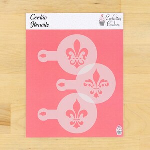May include: Pink cookie stencils with a fleur-de-lis design. The stencils are round with a handle and are arranged on a pink background. The words "Cookie Stencils" are printed at the top, and the Confection Couture logo is visible.