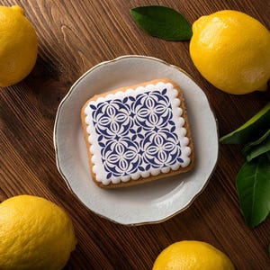 May include: A white cookie with a blue and white geometric pattern on top, sitting on a white plate. The cookie is surrounded by lemons on a wooden surface.