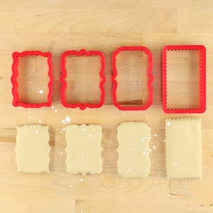 May include: Four red plastic cookie cutters in different shapes, with four pieces of cookie dough cut out using the cutters.