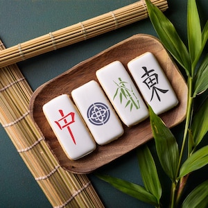 May include: Four white rectangular cookies decorated with Chinese characters and a bamboo design. The cookies are arranged on a wooden tray with a bamboo mat and green leaves in the background.