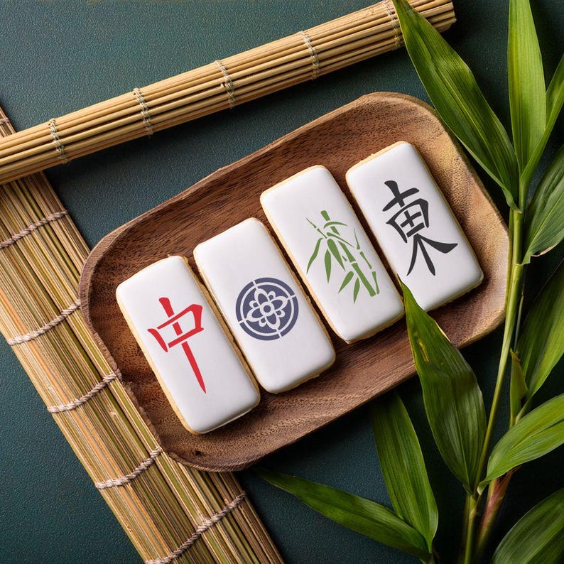May include: Four white rectangular cookies decorated with Chinese characters and a bamboo design. The cookies are arranged on a wooden tray with a bamboo mat and green leaves in the background.