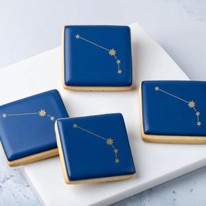 Könnte beinhalten: Vier quadratische Kekse mit einem goldenen Sternbild-Design auf einer glänzenden, dunkelblauen Oberfläche. Die Kekse haben eine hellbraune, rechteckige Basis. Die Kekse sind auf einer weißen Oberfläche angeordnet.