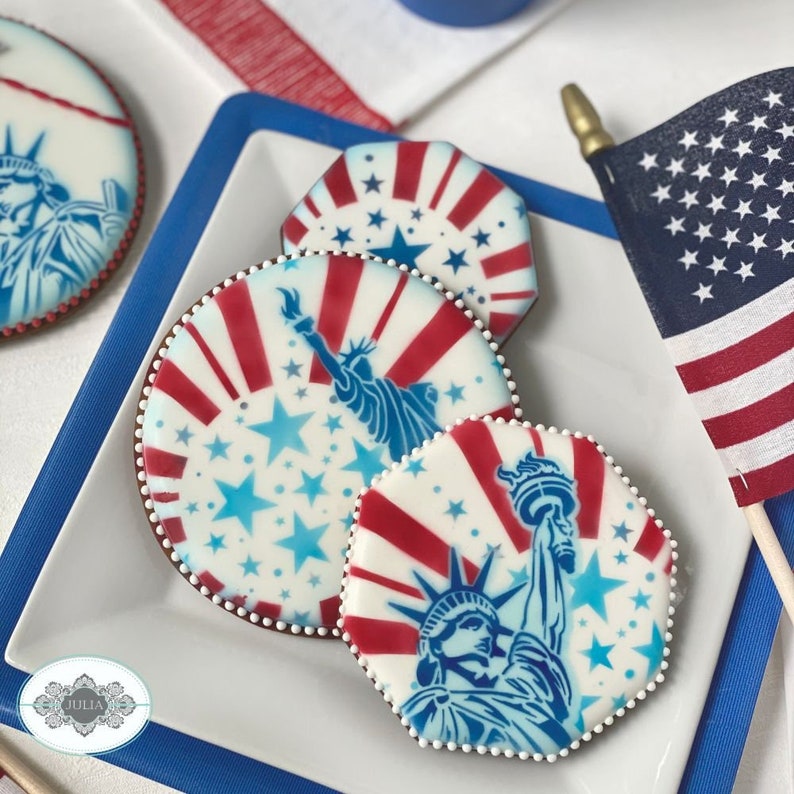 Puede incluir: Galletas decoradas con dise&ntilde;os patri&oacute;ticos que presentan la Estatua de la Libertad, rayas rojas y blancas y estrellas azules. Las galletas est&aacute;n dispuestas en un plato blanco con un borde azul, junto a una peque&ntilde;a bandera estadounidense.