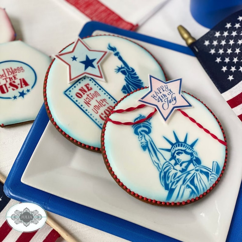 Puede incluir: Dos galletas redondas decoradas con temas patri&oacute;ticos. Una presenta la Estatua de la Libertad y el texto "Happy 4th of July" en una estrella. La otra galleta tiene la Estatua de la Libertad y el texto "One Nation Under God".