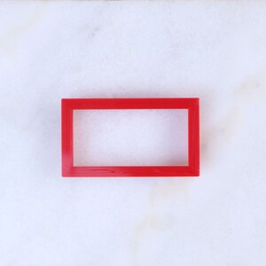 May include: A red plastic rectangular frame with a blank space inside. The frame is on a white marble surface.