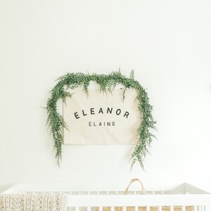 May include: A white cot with a white knitted blanket and a woven basket. A canvas sign with the text "Eleanor Elaine" is hanging above the cot, surrounded by a garland of green leaves.