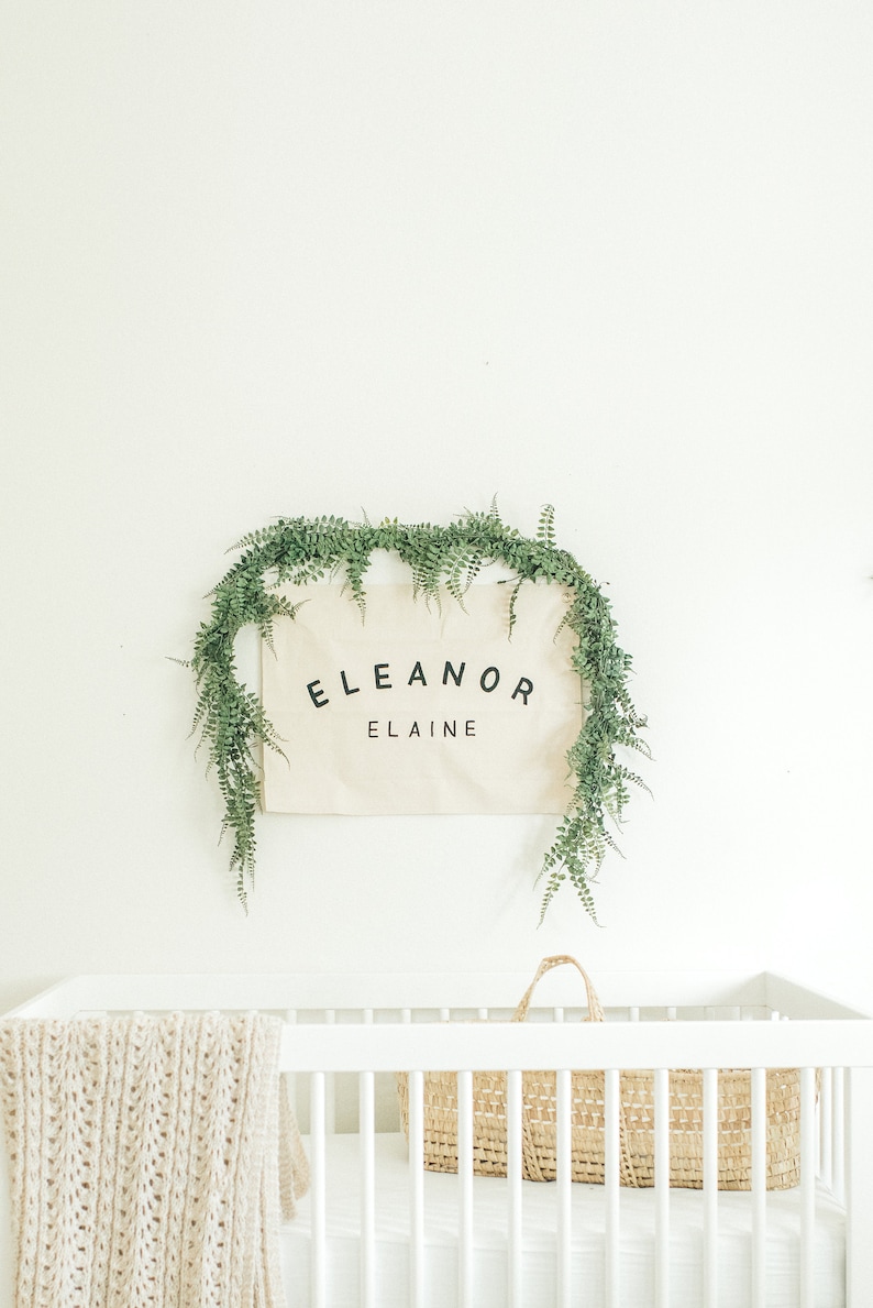 May include: A white cot with a white knitted blanket and a woven basket. A canvas sign with the text "Eleanor Elaine" is hanging above the cot, surrounded by a garland of green leaves.