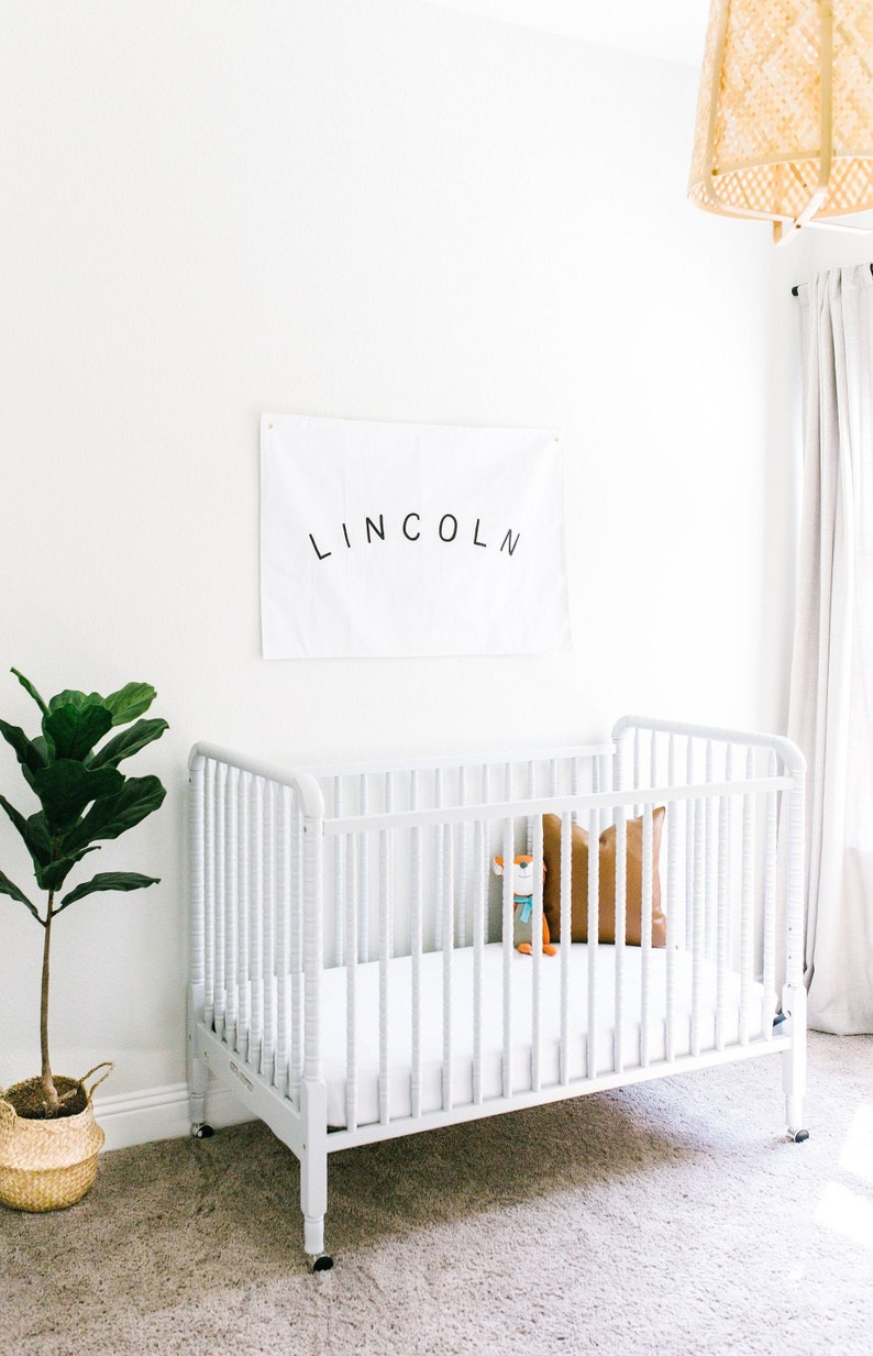May include: White cot with a white mattress and a brown pillow. A stuffed toy is in the cot. A white banner with the name "LINCOLN" is on the wall behind the cot.