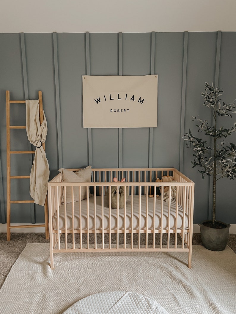 May include: A light wood cot with a white mattress and a white blanket. A small stuffed toy is in the cot. A canvas banner with the text "WILLIAM ROBERT" is hanging on the wall behind the cot. A small olive tree is in a pot on the right side of the image.