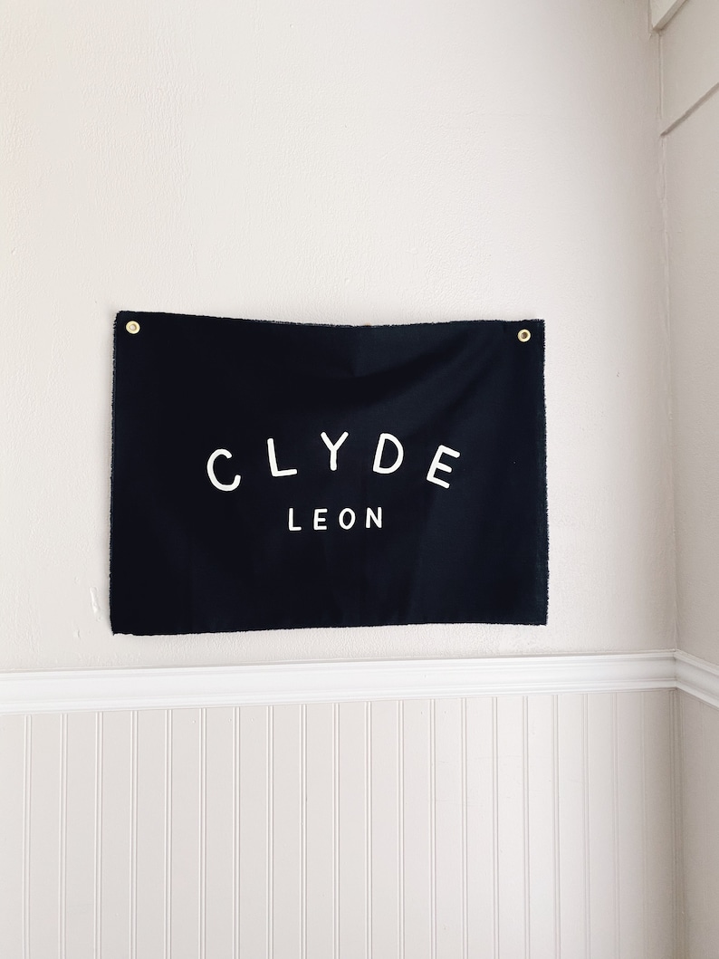 May include: Black fabric banner with white text that reads "CLYDE LEON" hanging on a white wall with white wainscoting.