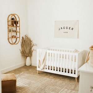 May include: A white cot with a white blanket and a pillow in a nursery with a beige rug, a wooden shelf with decorative items, and a white wall with a banner that reads "Jagger Luck".