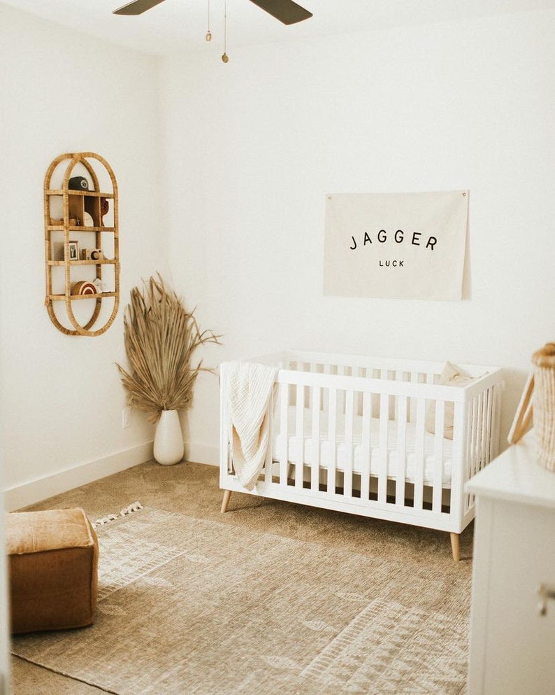 May include: A white cot with a white blanket and a pillow in a nursery with a beige rug, a wooden shelf with decorative items, and a white wall with a banner that reads "Jagger Luck".