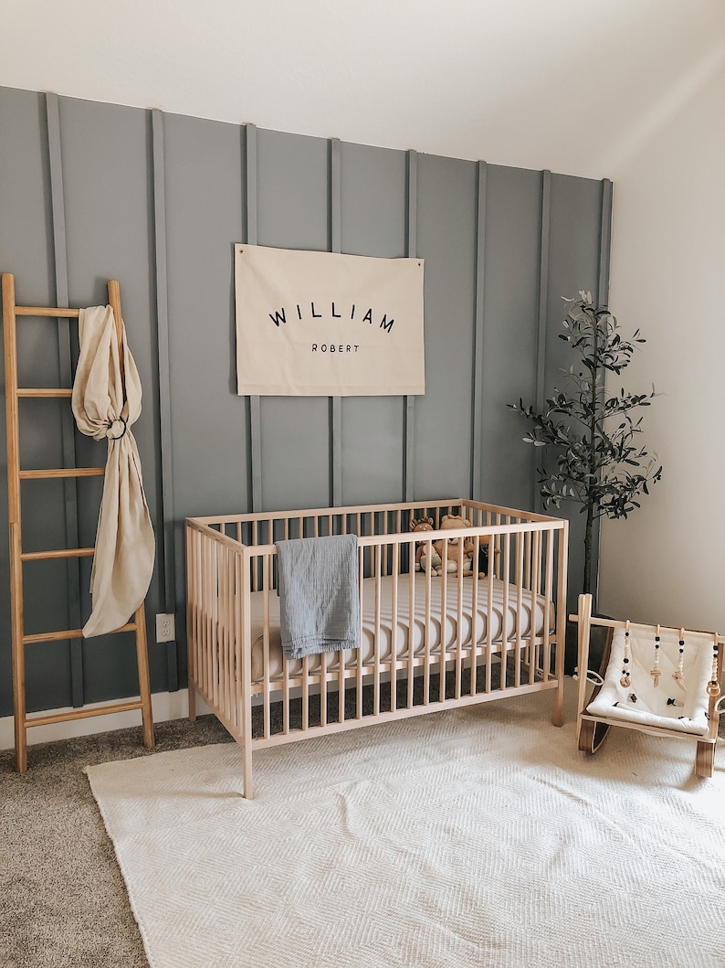 May include: A light wood cot with a white and grey striped mattress, a grey blanket, and a few stuffed toys. A wooden rocking chair with a beaded mobile hangs above it. A grey wall with wood panelling features a canvas banner with the text "WILLIAM ROBERT".