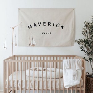 May include: A white cot with a white and beige blanket and a white pillow. A beige canvas banner with the text "Maverick Wayne" hangs on the wall above the cot.