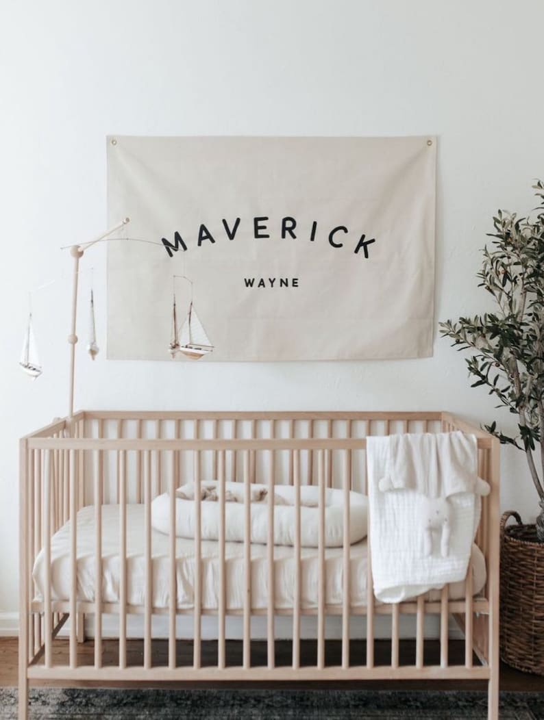 May include: A white cot with a white and beige blanket and a white pillow. A beige canvas banner with the text "Maverick Wayne" hangs on the wall above the cot.