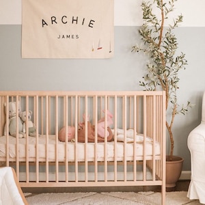 May include: A light wood cot with a baby lying on a white sheet. The cot is in a nursery with a white and light blue wall, a white rug, and a potted plant. A banner with the text "ARCHIE JAMES" hangs on the wall.