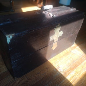 May include: A dark brown wooden chest with a metal latch and a brass decorative piece on the front. The chest is sitting on a wooden floor.