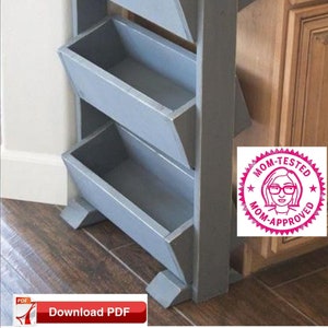 Puede incluir: Una estantería de madera de tres niveles con estantes pintados de gris. Los estantes están inclinados y tienen un aspecto rústico. La unidad está de pie en una cocina junto a un armario con una encimera de granito. La unidad tiene una pegatina "Probado por mamá, aprobado por mamá".