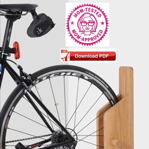 Puede incluir: Un soporte de pared de madera para bicicletas sostiene una rueda de bicicleta. La bicicleta es negra con una luz trasera roja y una bolsa de asiento negra. La rueda tiene neumáticos negros y radios plateados. Un sello rosa "Mom-Tested Mom-Approved" está en la parte superior derecha.