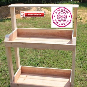 Puede incluir: Un soporte de plantas de madera de dos niveles con un acabado de madera natural. El soporte tiene un diseño sencillo con dos estantes rectangulares y una barandilla superior. El soporte está al aire libre sobre una superficie cubierta de hierba. Una pegatina rosa y blanca con el texto "Mom-Tested Mom-Approved" está pegada a la barandilla superior.