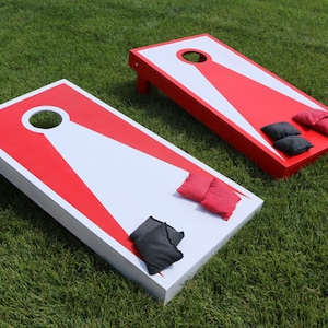 Può includere: Due tavole di cornhole in legno bianco e rosso con sacchi di fagioli neri. Le tavole hanno un triangolo bianco al centro con un cerchio rosso al centro del triangolo.