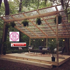 Puede incluir: Una pérgola de madera con un techo de plástico transparente, rodeada de árboles. La pérgola tiene una terraza de madera con asientos y una mesa. Hay plantas colgantes y luces de cadena.