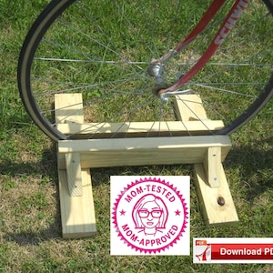 Puede incluir: Un soporte de bicicleta de madera con una rueda de bicicleta roja apoyada en él. El soporte está hecho de dos piezas de madera que están conectadas por una tercera pieza de madera. El soporte está sentado en un césped verde. El soporte tiene una pegatina rosa y blanca que dice "Probado por mamá Aprobado por mamá".
