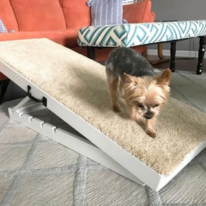 Op de afbeelding: Witte huisdierhelling met een beige tapijt oppervlak. Een kleine hond loopt de helling op.