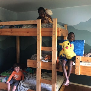 May include: Three children are on a wooden bunk bed with a mountain mural on the wall behind them. The top bunk has a stuffed giraffe and a pillow. The middle bunk has a stuffed animal and a blanket. The bottom bunk has a blanket and a pillow.