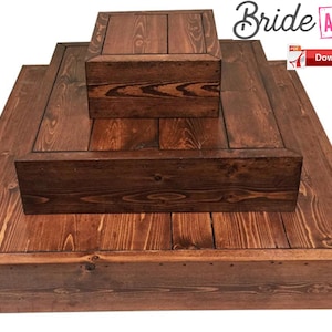 Könnte beinhalten: Ein dreistufiger Kuchenständer aus Holz mit rustikalem braunen Finish. Der Ständer ist aus Naturholz mit quadratischer Form und schlichtem Design. Die oberste Stufe ist die kleinste und die unterste Stufe ist die größte. Der Ständer ist perfekt zum Präsentieren von Kuchen, Cupcakes oder anderen Desserts.