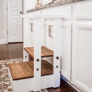 Puede incluir: Taburete de madera blanco con dos pelda&ntilde;os de madera. El taburete est&aacute; delante de armarios de cocina blancos.