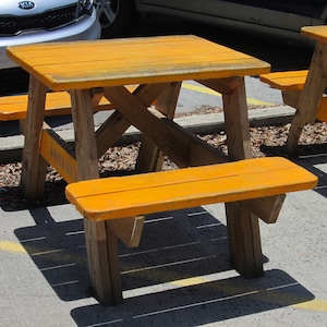 Könnte beinhalten: Ein hölzerner Picknicktisch mit zwei Bänken. Tisch und Bänke sind in einem leuchtenden Orange gestrichen. Der Tisch steht vor einem Auto.