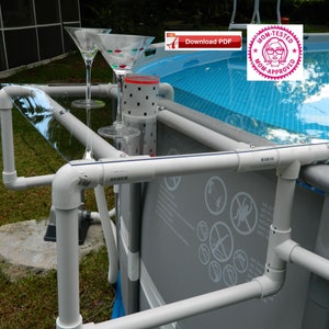 May include: A white PVC pipe poolside table with a clear glass top. Two martini glasses are on the table. The table is attached to the side of a blue and gray above ground pool.