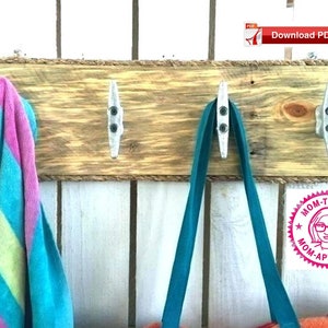 Może przedstawiać: Drewniany stojak na ubrania montowany na ścianie z ozdobną linką i trzema metalowymi haczykami w kształcie cum. Ręcznik w paski niebiesko-różowe wisi na jednym haczyku, a turkusowa torba na ramię na drugim. Na różowej okrągłej naklejce widnieje napis "Mom-Tested Mom-Approved".