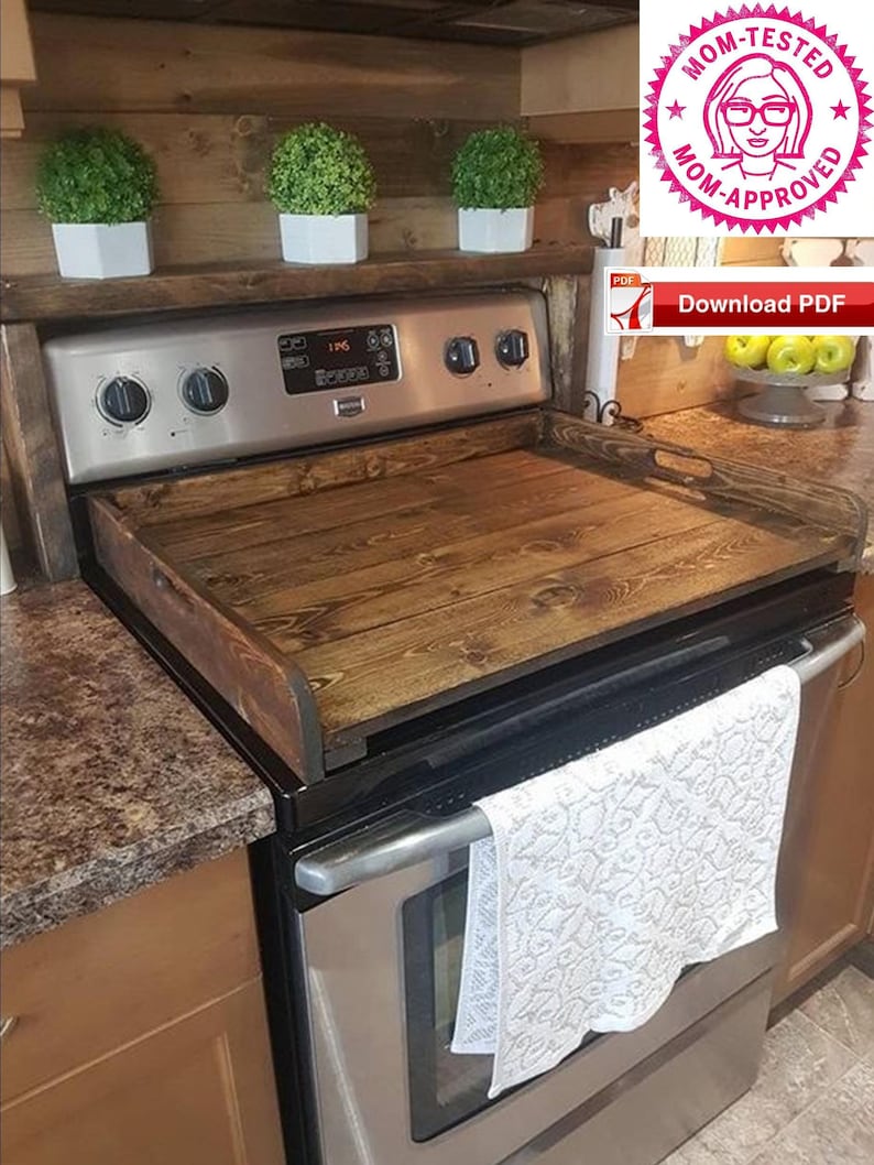 May include: A wooden stovetop cover with a dark brown stain. The cover is placed on top of a stainless steel stove with a white towel hanging on the oven door.