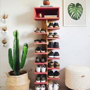 May include: A red and wood shoe rack with eight shelves, filled with various shoes. The rack has a small shelf on top with a gold lamp and books. A cactus in a woven basket is on the left, and a white crocheted ottoman is on the right.