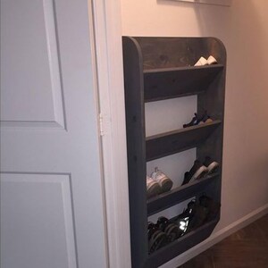 K&ouml;nnte beinhalten: Ein dunkelgrauer Schuhschrank aus Holz mit f&uuml;nf Regalen, der an einer Wand montiert ist. Der Schuhschrank ist abgewinkelt und hat abgerundete Kanten. Die Regale sind mit Schuhen gef&uuml;llt.