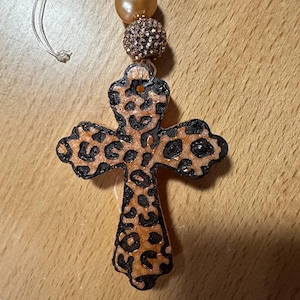 May include: A cross-shaped ornament with a leopard print design in shades of brown and black. The ornament is suspended by a clear string and adorned with two decorative beads in gold and bronze tones. The cross is likely a religious item.