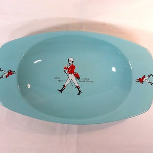 May include: A light blue ceramic ashtray with a walking man in a red jacket and white pants. The text "BORN 1820 - STILL GOING STRONG" is printed on the ashtray.