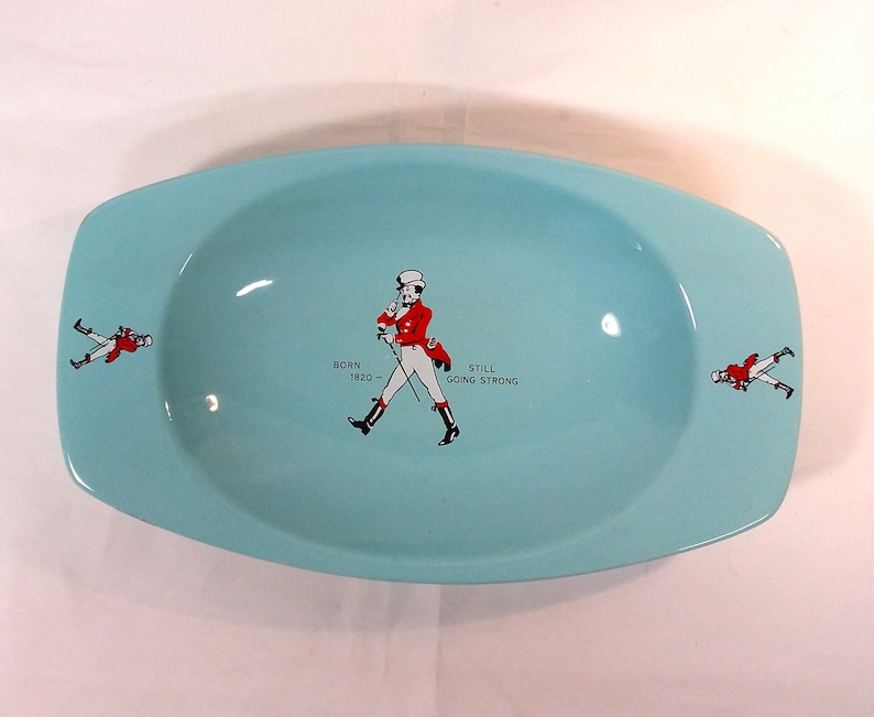 May include: A light blue ceramic ashtray with a walking man in a red jacket and white pants. The text "BORN 1820 - STILL GOING STRONG" is printed on the ashtray.
