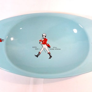 May include: A light blue oval-shaped ceramic ashtray with a Johnnie Walker logo. The logo features a man in a red coat and white pants walking with a cane. The text "Born 1820 - Still Going Strong" is printed below the man.