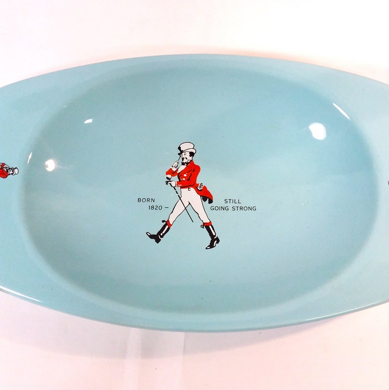 May include: A light blue oval-shaped ceramic ashtray with a Johnnie Walker logo. The logo features a man in a red coat and white pants walking with a cane. The text "Born 1820 - Still Going Strong" is printed below the man.