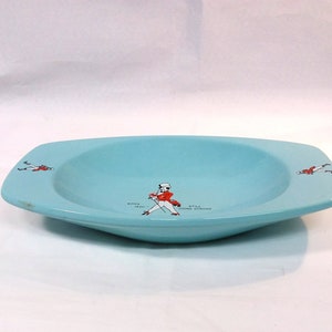 May include: A light blue ceramic dish with a rounded bottom and squared edges. The dish has a white and red illustration of a man in a red jacket and black pants walking with a cane. The text "BORN 1820 STILL GOING STRONG" is printed on the dish.