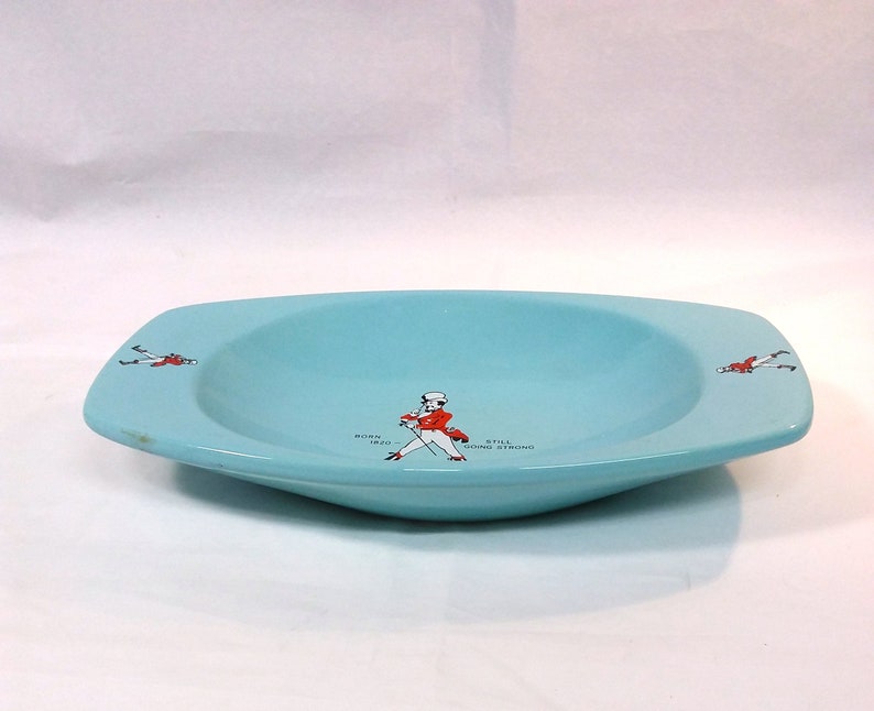 May include: A light blue ceramic dish with a rounded bottom and squared edges. The dish has a white and red illustration of a man in a red jacket and black pants walking with a cane. The text "BORN 1820 STILL GOING STRONG" is printed on the dish.