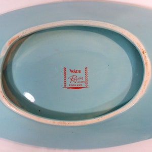May include: A light blue oval ceramic dish with a white rim. The bottom of the dish has a red logo that reads "Wade Regalo London England" with a wreath on either side of the text.